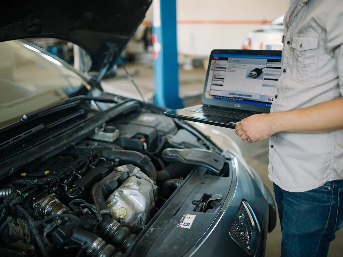 Technician holding a laptop beside an open car hood during an inspection